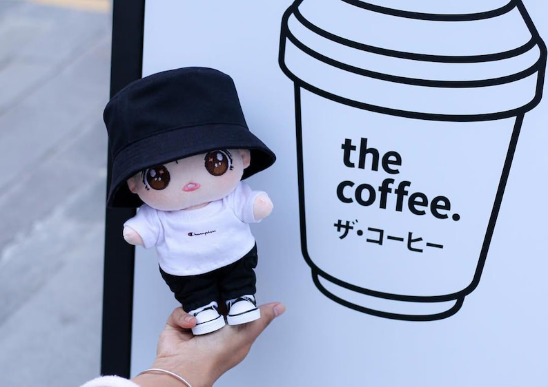 May include: A plush doll wearing a black bucket hat, white t-shirt, black pants, and sneakers. The doll is next to a sign with a coffee cup graphic and the words "the coffee." and Japanese text.