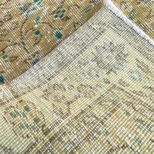 May include: Close-up of a section of a vintage rug with a distressed, faded appearance. The rug features a beige and gold color palette with teal accents. The design includes floral and geometric patterns, showcasing a traditional style.