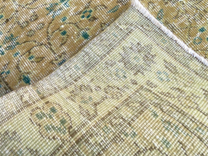 May include: Close-up of a section of a vintage rug with a distressed, faded appearance. The rug features a beige and gold color palette with teal accents. The design includes floral and geometric patterns, showcasing a traditional style.
