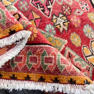May include: A hand-knotted wool rug featuring a vibrant red base with intricate geometric patterns in yellow, green, pink, and black. The rug has a thick, textured pile and is finished with white knotted fringe along the edges, showing a folded corner.