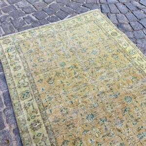May include: A rectangular, vintage-style rug with a faded, distressed appearance. The rug features a central field of muted yellow and beige tones, adorned with floral and botanical patterns in shades of teal and brown. The border is a lighter green.