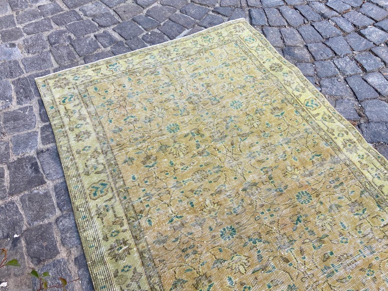 May include: A rectangular, vintage-style rug with a faded, distressed appearance. The rug features a central field of muted yellow and beige tones, adorned with floral and botanical patterns in shades of teal and brown. The border is a lighter green.