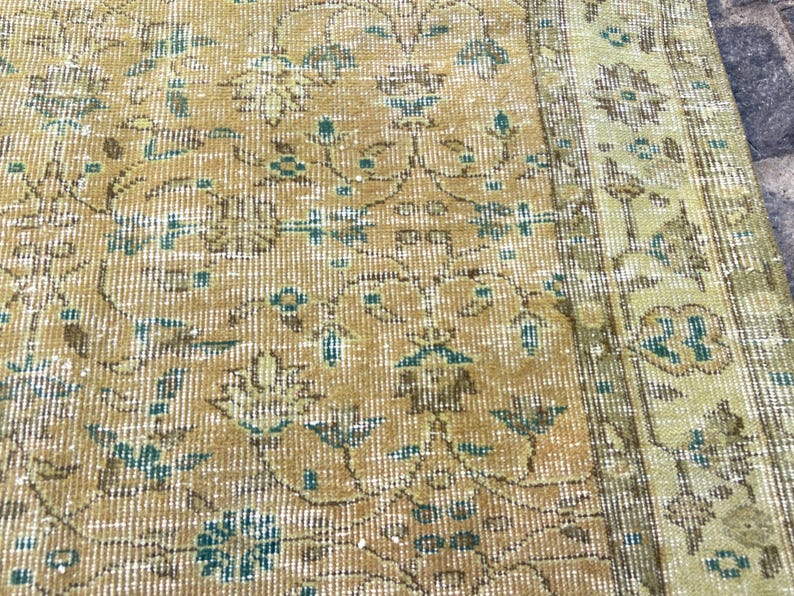 May include: Close-up of a vintage rug with a distressed, floral pattern. The rug features a warm color palette of gold, beige, and teal. The design includes stylized flowers and vines, with a faded, antique appearance.