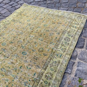 May include: An antique, rectangular rug with a distressed, floral pattern. The rug features a warm, golden-yellow field with faded green and blue accents. The border is a lighter green with a similar floral design. The rug is placed on a cobblestone surface.