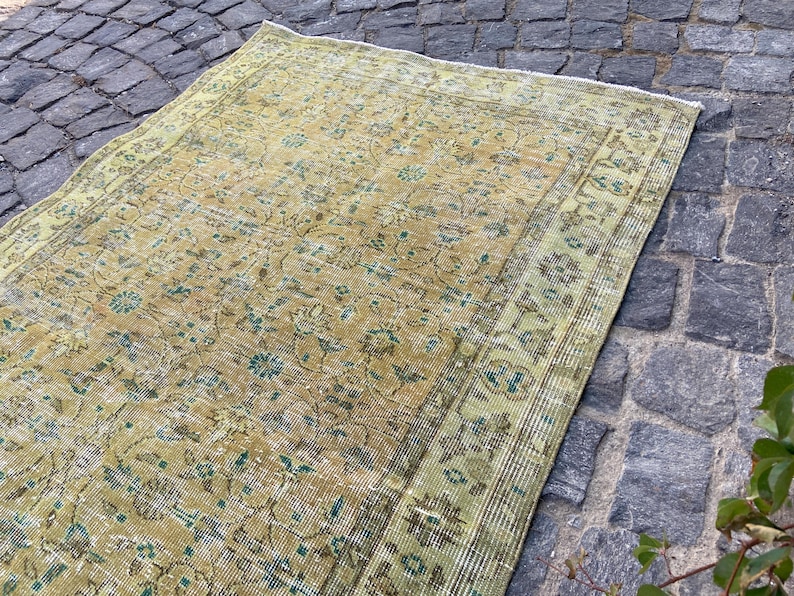May include: An antique, rectangular rug with a distressed, floral pattern. The rug features a warm, golden-yellow field with faded green and blue accents. The border is a lighter green with a similar floral design. The rug is placed on a cobblestone surface.