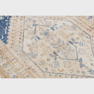 May include: Close-up of a beige and blue patterned rug. The rug features a central diamond design with floral and geometric motifs in shades of blue and tan. The border has a dark blue geometric pattern.