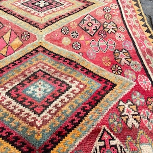 May include: A woven rug featuring a repeating diamond pattern in shades of red, pink, yellow, teal, and black. The design includes intricate geometric motifs and small floral-like shapes within the diamonds, finished with a decorative border.