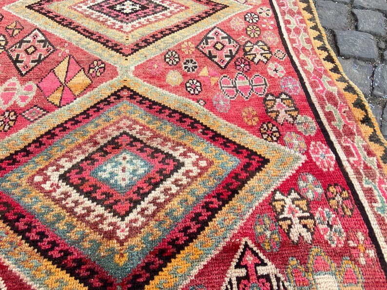 May include: A woven rug featuring a repeating diamond pattern in shades of red, pink, yellow, teal, and black. The design includes intricate geometric motifs and small floral-like shapes within the diamonds, finished with a decorative border.