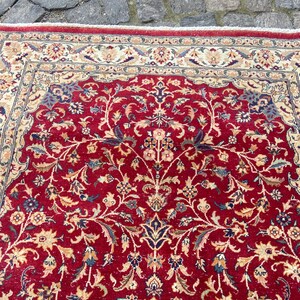 May include: A red rug with a floral pattern. The rug has a cream-colored border with a dark blue and cream-colored inner border.