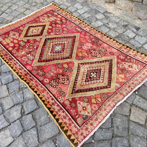 May include: A rectangular area rug featuring a vibrant red base color with three large central diamond motifs in shades of beige, green, and black. The rug is decorated with intricate geometric patterns and has a dark border with fringe edges.