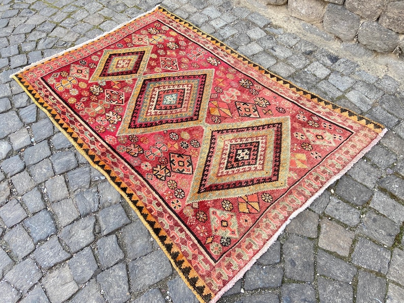 May include: A rectangular area rug featuring a vibrant red base color with three large central diamond motifs in shades of beige, green, and black. The rug is decorated with intricate geometric patterns and has a dark border with fringe edges.