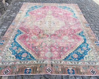 May include: Large, rectangular, vintage rug with a central pink field and intricate geometric patterns. The rug features a teal blue border and accents, with a brown and beige outer border. The rug is placed on a stone surface.