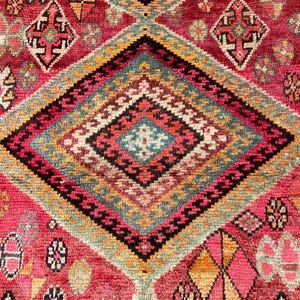 May include: A woven rug featuring a vibrant red background with a large central diamond motif. The pattern includes intricate geometric shapes in shades of teal, yellow, black, and white, with smaller repeating floral and diamond accents throughout.