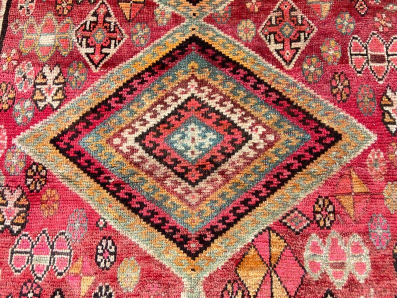 May include: A woven rug featuring a vibrant red background with a large central diamond motif. The pattern includes intricate geometric shapes in shades of teal, yellow, black, and white, with smaller repeating floral and diamond accents throughout.