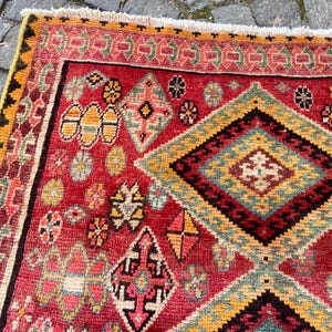 May include: A rectangular rug featuring a vibrant red field decorated with intricate geometric patterns, including large diamonds, smaller floral motifs, and serrated borders in shades of yellow, green, black, and pink, finished with a white fringe.