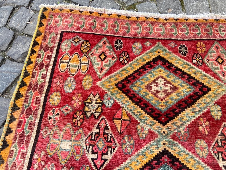 May include: A rectangular rug featuring a vibrant red field decorated with intricate geometric patterns, including large diamonds, smaller floral motifs, and serrated borders in shades of yellow, green, black, and pink, finished with a white fringe.