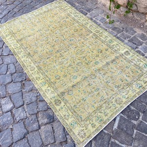 May include: Rectangular, vintage-style rug with a faded, floral pattern in shades of yellow, green, and brown. The rug has a detailed border and is placed on a cobblestone surface. The overall design suggests a traditional, antique aesthetic.