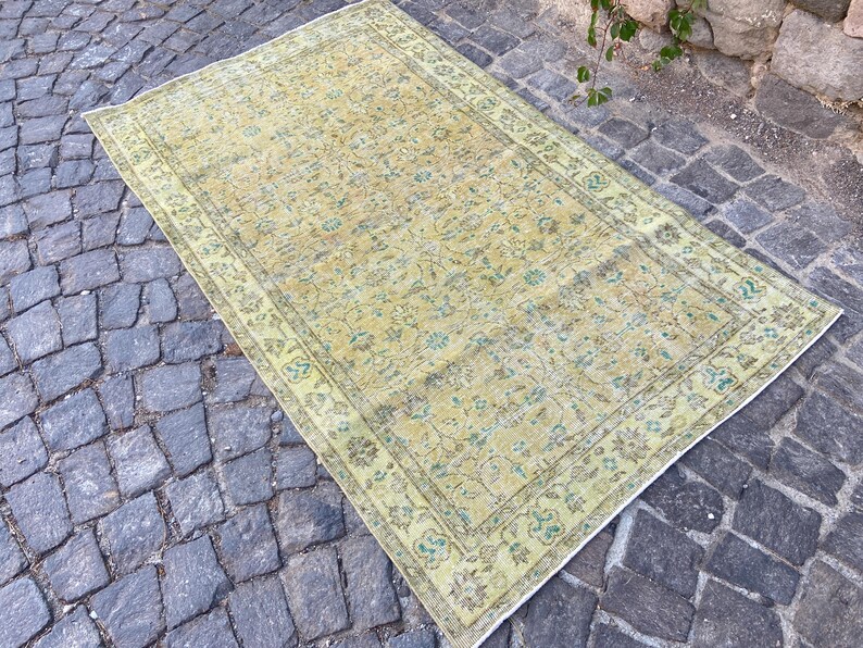 May include: Rectangular, vintage-style rug with a faded, floral pattern in shades of yellow, green, and brown. The rug has a detailed border and is placed on a cobblestone surface. The overall design suggests a traditional, antique aesthetic.