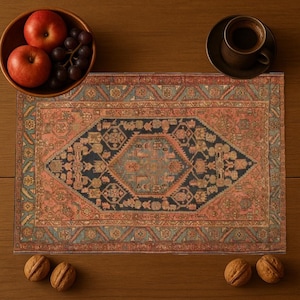 May include: An overhead shot of a rectangular rug with a central navy blue diamond design and a coral border. A wooden bowl of red apples and grapes, a cup of coffee, and walnuts are arranged around the rug on a wooden surface.