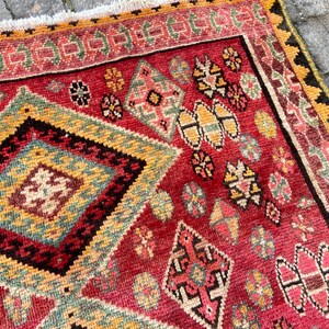 May include: A rectangular woven rug featuring a vibrant red background with a large central diamond pattern in yellow, teal, and black. The rug is decorated with smaller geometric motifs in pink, green, and yellow, finished with a white fringe border.