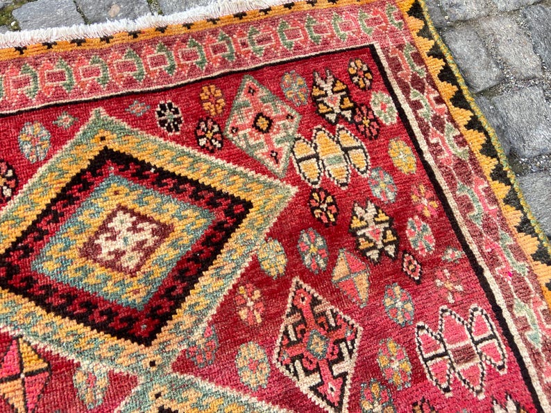 May include: A rectangular woven rug featuring a vibrant red background with a large central diamond pattern in yellow, teal, and black. The rug is decorated with smaller geometric motifs in pink, green, and yellow, finished with a white fringe border.