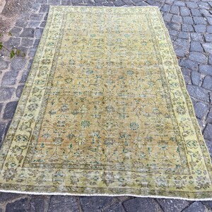 May include: An antique, rectangular rug with a distressed, faded appearance. The rug features a central field of muted yellow-green with floral patterns and a border in a similar color. The rug is placed on a cobblestone surface.