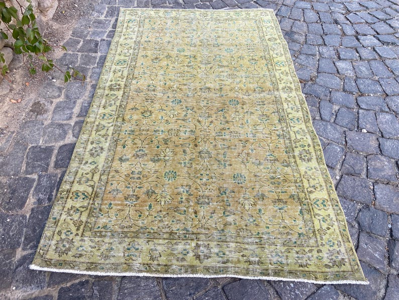 May include: An antique, rectangular rug with a distressed, faded appearance. The rug features a central field of muted yellow-green with floral patterns and a border in a similar color. The rug is placed on a cobblestone surface.