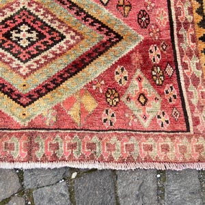 May include: A woven rug featuring a large central diamond pattern in shades of red, orange, green, and black. The surface displays intricate geometric motifs, small floral-like shapes, and a fringed edge, showcasing a dense, textured pile construction.