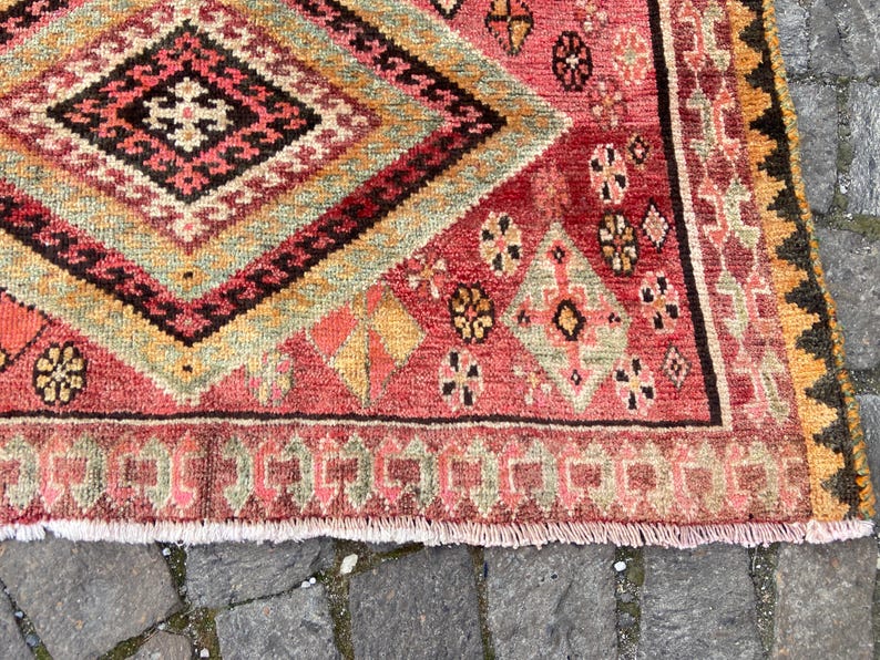 May include: A woven rug featuring a large central diamond pattern in shades of red, orange, green, and black. The surface displays intricate geometric motifs, small floral-like shapes, and a fringed edge, showcasing a dense, textured pile construction.
