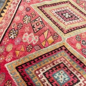 May include: A woven rug featuring a vibrant red base with large diamond-shaped patterns in shades of pink, yellow, blue, and black. The surface is decorated with intricate geometric motifs and small floral-like shapes throughout the design.