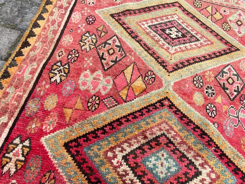 May include: A woven rug featuring a vibrant red base with large diamond-shaped patterns in shades of pink, yellow, blue, and black. The surface is decorated with intricate geometric motifs and small floral-like shapes throughout the design.