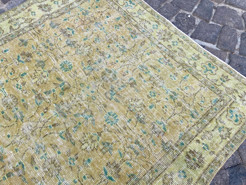 May include: An antique rug with a distressed, faded appearance. The rug features a repeating floral pattern in shades of olive green, brown, and teal. The rug's design has a vintage aesthetic, suitable for home decor.