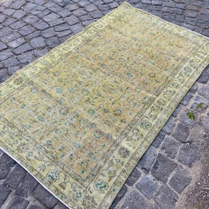 May include: A rectangular, vintage-style rug with a faded, distressed appearance. The rug features a central field of muted yellow and beige tones, with a detailed floral pattern. The border is a lighter shade of green with floral motifs.