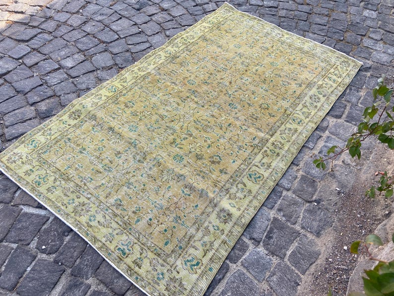 May include: A rectangular, vintage-style rug with a faded, distressed appearance. The rug features a central field of muted yellow and beige tones, with a detailed floral pattern. The border is a lighter shade of green with floral motifs.