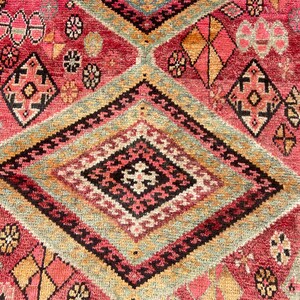 May include: A woven rug featuring a vibrant red background with large, repeating diamond patterns in shades of sage green, black, and cream. The design includes intricate geometric motifs, small floral-like shapes, and orange accents throughout.