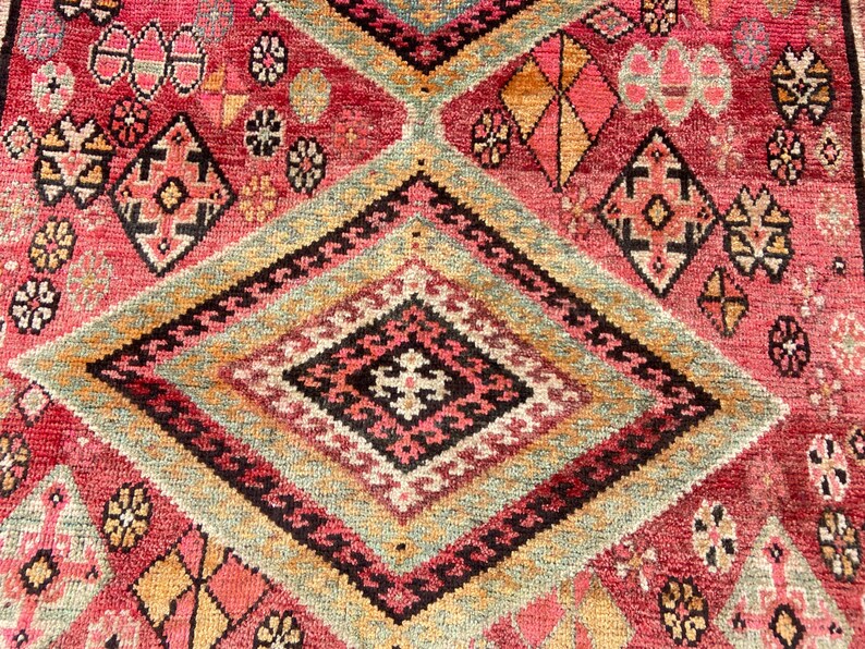 May include: A woven rug featuring a vibrant red background with large, repeating diamond patterns in shades of sage green, black, and cream. The design includes intricate geometric motifs, small floral-like shapes, and orange accents throughout.