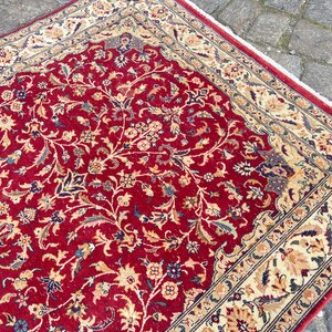 May include: A red rug with a floral pattern and a cream-colored border. The rug has a traditional design with intricate details.
