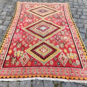 May include: A rectangular area rug featuring a vibrant red background with three large, centered diamond motifs in shades of yellow, green, and black. The rug is decorated with various small geometric patterns and has a fringed edge on the bottom.
