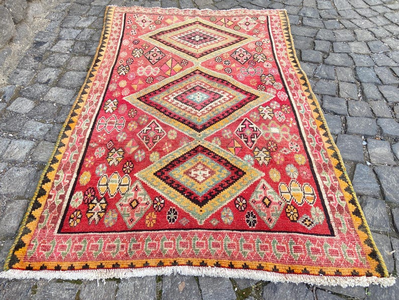 May include: A rectangular area rug featuring a vibrant red background with three large, centered diamond motifs in shades of yellow, green, and black. The rug is decorated with various small geometric patterns and has a fringed edge on the bottom.