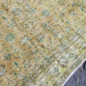 May include: Close-up of a vintage rug with a distressed, textured appearance. The rug features a warm color palette of gold, brown, and green, with intricate floral and geometric patterns. The rug's design includes teal accents.