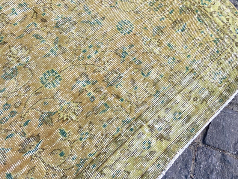 May include: Close-up of a vintage rug with a distressed, textured appearance. The rug features a warm color palette of gold, brown, and green, with intricate floral and geometric patterns. The rug's design includes teal accents.