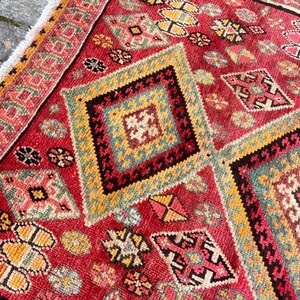 May include: A rectangular woven rug featuring a vibrant red background with repeating diamond-shaped geometric patterns in yellow, teal, black, and white. The rug includes a decorative border and white fringe along the edge of the textile.