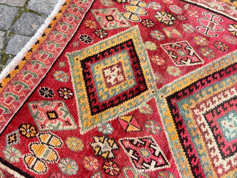 May include: A rectangular woven rug featuring a vibrant red background with repeating diamond-shaped geometric patterns in yellow, teal, black, and white. The rug includes a decorative border and white fringe along the edge of the textile.