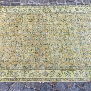 May include: A rectangular, vintage-style rug with a faded, distressed appearance. The rug features a central field of muted yellow and brown tones with floral and vine patterns. The border is a lighter green with similar floral motifs.