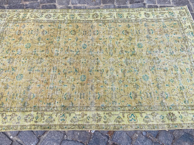 May include: A rectangular, vintage-style rug with a faded, distressed appearance. The rug features a central field of muted yellow and brown tones with floral and vine patterns. The border is a lighter green with similar floral motifs.