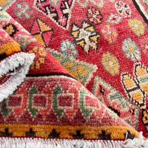 May include: A woven rug featuring a vibrant red base with intricate geometric patterns in yellow, green, and black. The rug has a folded corner revealing a white fringed edge along the border, showcasing a dense, textured pile construction.