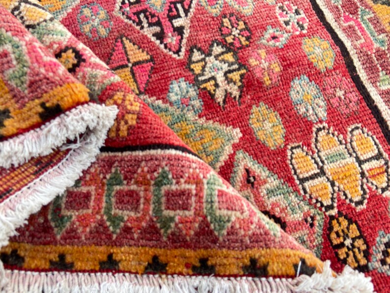 May include: A woven rug featuring a vibrant red base with intricate geometric patterns in yellow, green, and black. The rug has a folded corner revealing a white fringed edge along the border, showcasing a dense, textured pile construction.