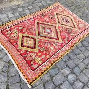 May include: A rectangular area rug featuring a vibrant red background with three prominent central diamond motifs in shades of yellow, green, and black. The rug is decorated with intricate geometric patterns and small floral accents, finished with a white fringe.