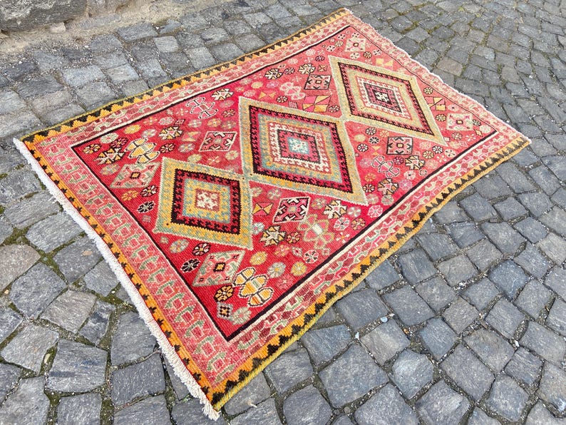 May include: A rectangular area rug featuring a vibrant red background with three prominent central diamond motifs in shades of yellow, green, and black. The rug is decorated with intricate geometric patterns and small floral accents, finished with a white fringe.