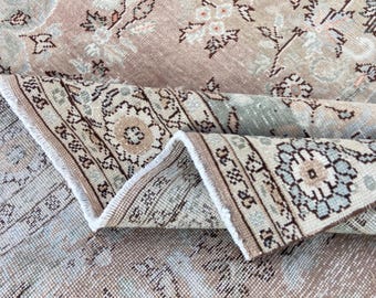 May include: A woven area rug featuring a muted tan and beige base with intricate floral patterns in shades of light blue, cream, and dark brown. The rug is folded over to reveal a detailed border with geometric motifs and a white fringe edge.